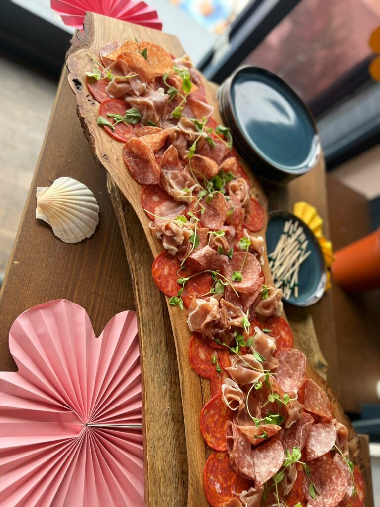 Large handmade wooden board with a mix of cheese and cold meats for sharing. Pink flowers and shells as decorations